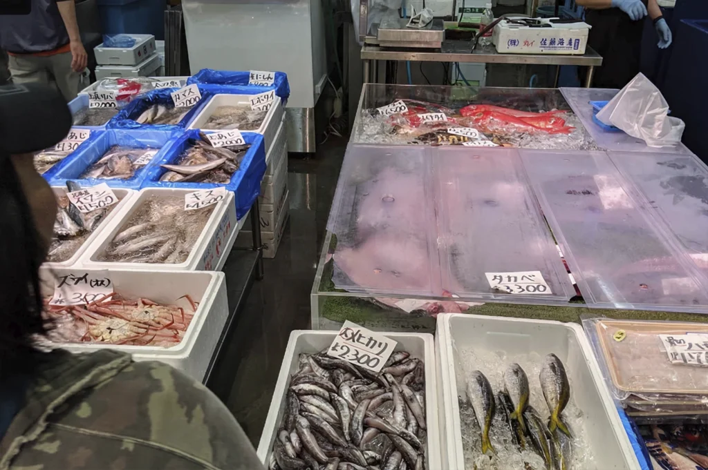 A high-angle, wide shot of a bustling Japanese fish market. Various types of fresh fish, crab, and seafood are displayed in white styrofoam and blue plastic bins filled with ice. Handwritten price tags in Japanese yen are tucked into each container. A person in a camouflage jacket is partially visible in the foreground, looking at the display.