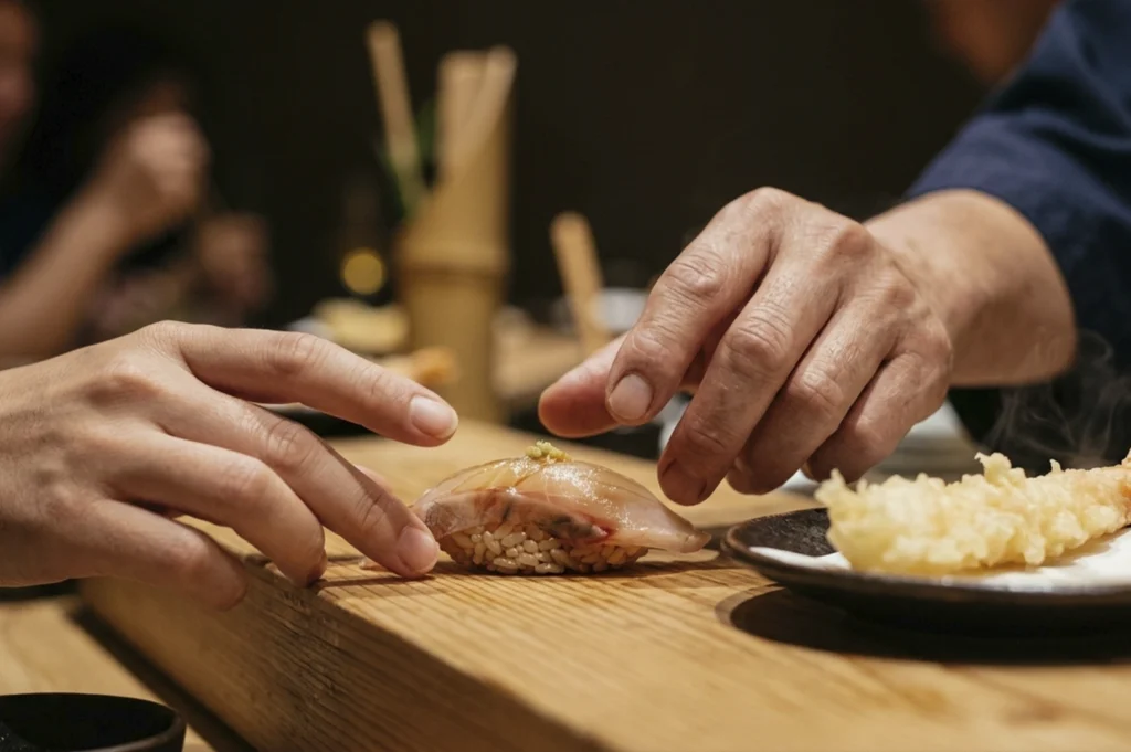 A close-up, side-profile shot of a piece of white fish nigiri topped with a tiny garnish. A diner’s hands are reaching out to pick up the sushi directly from the wooden counter using their fingers—a traditional way to eat nigiri. To the right, a small plate with a golden, crispy piece of shrimp tempura is partially visible. The lighting is warm and directional, highlighting the textures of the food and the wood.