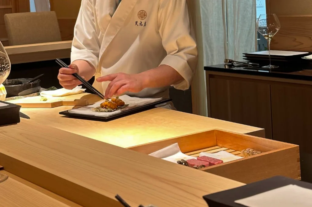 A medium shot of a chef dressed in a traditional white uwagi (chef's jacket) meticulously preparing a dish. Using long metal cooking chopsticks, the chef is placing a bright orange sea urchin (uni) garnish onto a small plate. In the foreground, a wooden box contains raw cuts of marbled wagyu beef, emphasizing the variety of premium ingredients used in a Kappo multi-course meal.