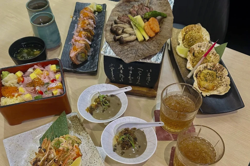 An overhead view of a wooden table showcases an extensive Japanese feast, featuring a colorful Chirashi bowl topped with cubed sashimi and ikura alongside a long platter of sushi rolls finished with shaved onions. In the center, a small charcoal shichirin grill cooks sliced beef and vegetables on a traditional magnolia leaf, flanked by a plate of three grilled scallops in their shells and two small bowls containing a creamy, greyish-brown appetizer. The meal is completed with two glasses of iced tea, bowls of miso soup, and a side of grilled prawns, all served on rustic ceramic dinnerware.