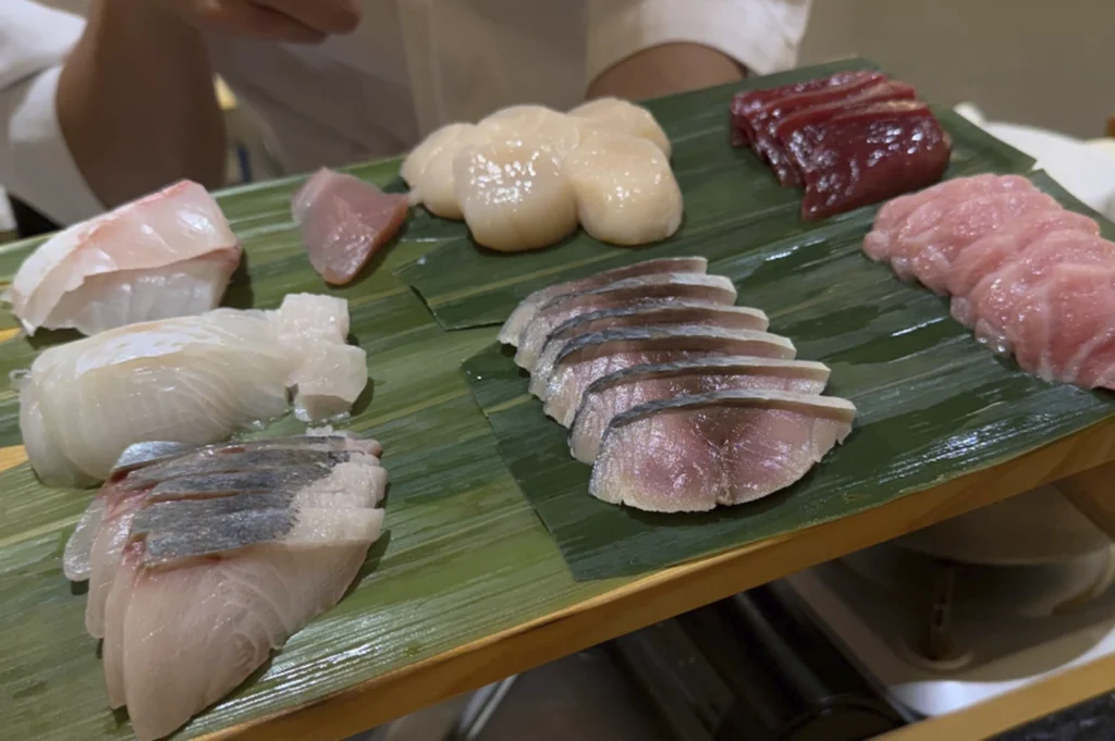 A wooden tray lined with green bamboo leaves showcases a variety of fresh, raw seafood prepared for sushi. The selection includes plump white scallops, translucent slices of white fish (tai or hirame), silver-skinned mackerel (shime saba), and various cuts of tuna ranging from lean red to fatty pink otoro. The chefs' white uniforms are visible in the soft-focus background.