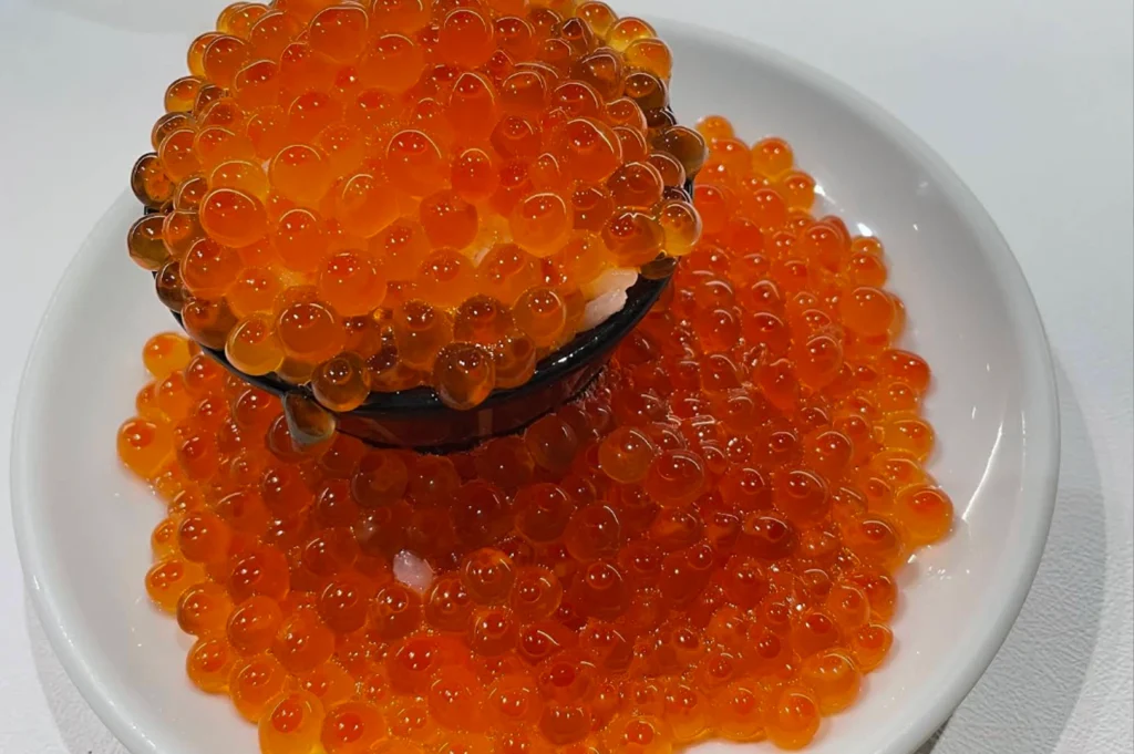 A close-up, slightly high-angle shot captures a mountain of bright, translucent orange salmon roe, or ikura, overflowing from a small, round black bowl. The bowl sits at the center of a shallow white plate, completely overwhelmed by the abundance of glistening, spherical eggs that cascade over its edges and pool thickly onto the plate below. Under bright, clean lighting, the ikura gems appear plump and vibrant, with subtle red-orange cores visible inside each sphere. A few grains of white sushi rice peek through the gaps just beneath the top layer of roe inside the bowl, emphasizing the extreme proportion of the topping. The stark white background and plate contrast sharply with the deep black of the bowl and the intense, warm orange hues of the seafood.