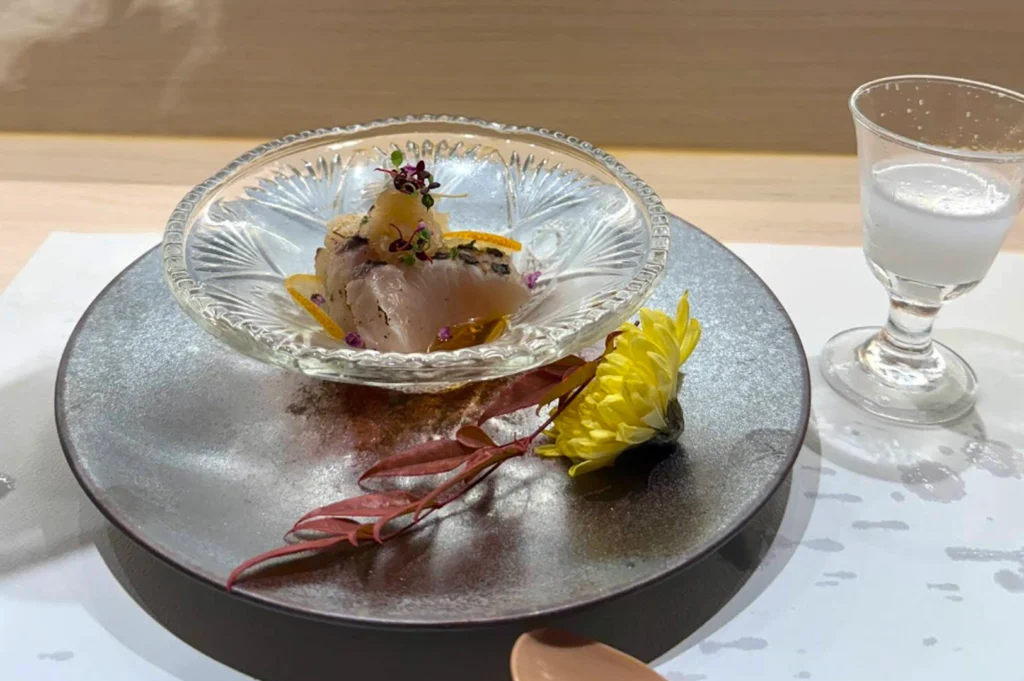 A sophisticated culinary shot of a small seafood dish served in a delicate, patterned clear glass bowl. The bowl contains pieces of sliced fish topped with grated daikon, microgreens, and citrus zest, sitting in a shallow pool of light sauce. The glass bowl is centered on a large, matte charcoal-grey plate, garnished with a dried reddish leaf and a yellow flower. To the right, a small glass of cloudy white liquid, possibly nigori sake, sits on the white table runner. The background shows the clean lines of a light wood counter.