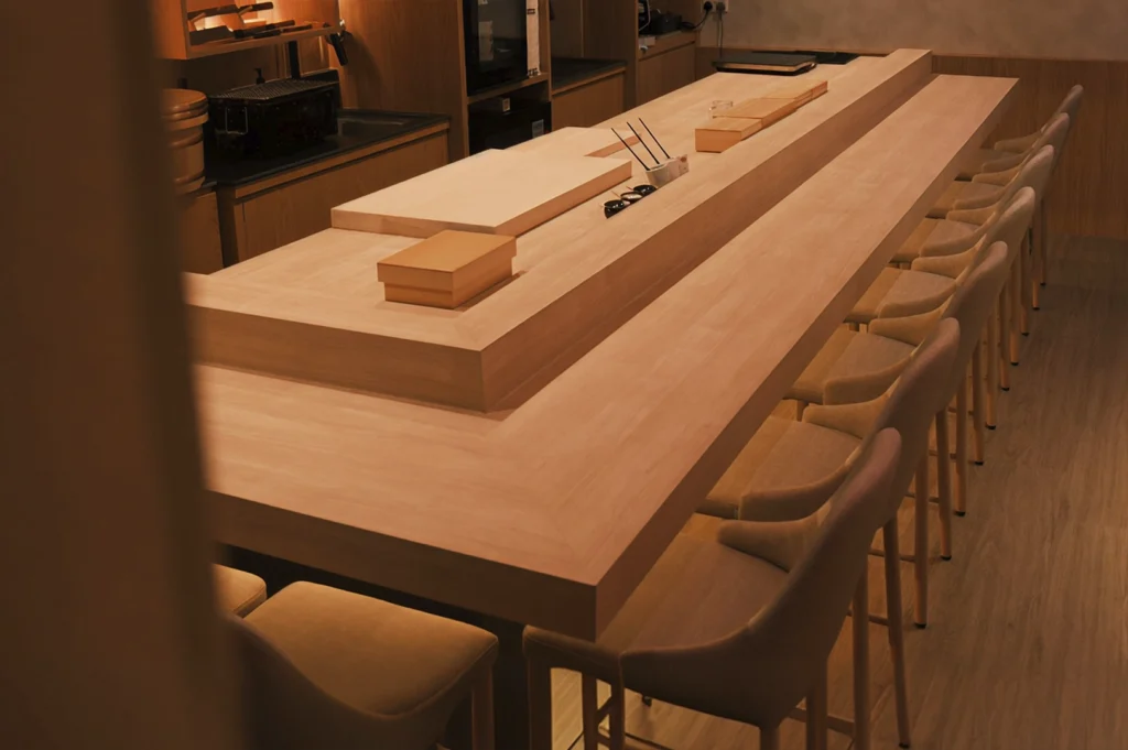 A wide interior shot of an empty, high-end sushi omakase counter made of light-colored Hinoki wood. The L-shaped wooden bar is lined with modern, minimalist chairs. The background features a clean, organized prep area with traditional Japanese knives and wooden containers under warm, ambient lighting.