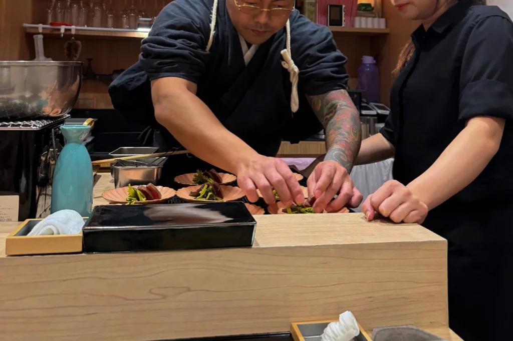 A medium shot captures a sushi chef with gold-rimmed glasses and a colorful floral sleeve tattoo on his left arm as he carefully plates small appetizers, assisted by a female staff member on the right. The chef, dressed in a dark blue wrap uniform, leans over the kitchen workstation to arrange delicate green garnishes onto several small, flower-shaped ceramic dishes holding sliced fish. In the foreground, the clean lines of a light-colored wooden counter are visible, adorned with a glossy black serving box, a light-blue sake carafe, and small wooden boxes containing wet hand towels.