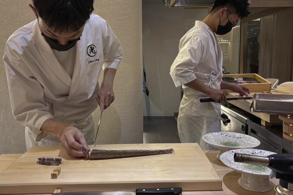 Two sushi chefs wearing white tunics and black face masks work behind a light wood prep counter. The chef in the foreground is meticulously slicing a long, silver-skinned fish with a long, thin yanagiba knife. To his right, the second chef prepares smaller garnishes. On the counter sit white footed serving plates and a culinary blowtorch, highlighting the blend of traditional knife skills and modern techniques.