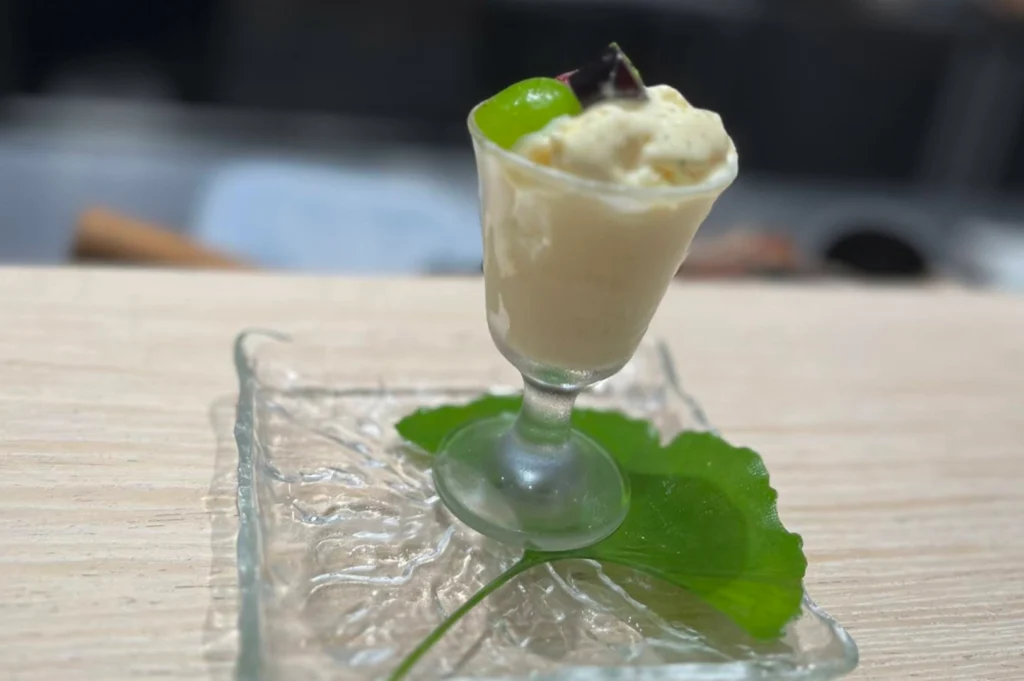 A close-up shot of a dessert served in a small, stemmed glass. The glass contains a creamy, pale yellow mousse or ice cream, topped with a bright green grape and a dark garnish. The glass sits on a square, textured clear glass coaster, which is placed over a vibrant green ginkgo leaf. The presentation is set on a light-toned wooden surface with a shallow depth of field, blurring the background into soft neutral tones.