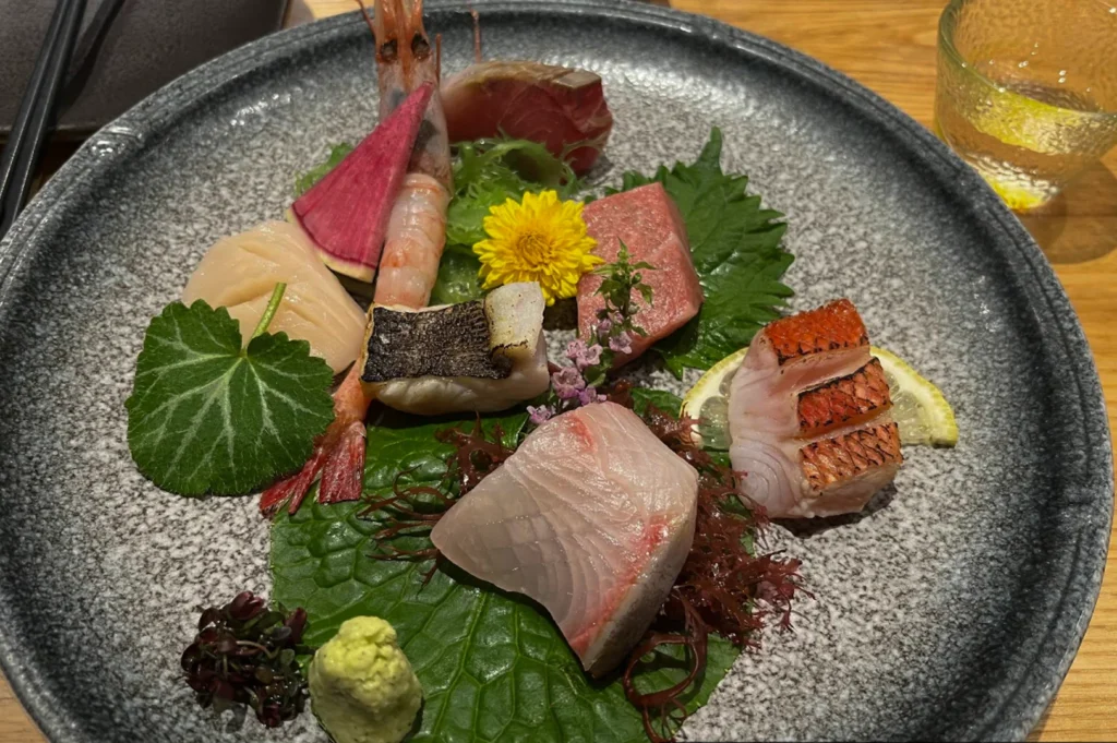 A colorful sashimi assortment served on a large, grey speckled ceramic plate, including slices of various raw fish, a raw shrimp, scallops, and garnishes such as a yellow chrysanthemum, shiso leaf, radish slice, and a dollop of fresh wasabi.