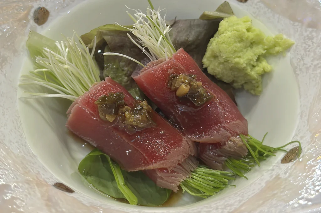 A top-down close-up of a Japanese appetizer served in a textured glass bowl. Two rolls of deep red raw tuna are stuffed with bright green sprouts and topped with a savory garnish. A generous dollop of fresh, pale green wasabi sits to the side on a bed of dark garnish leaves.