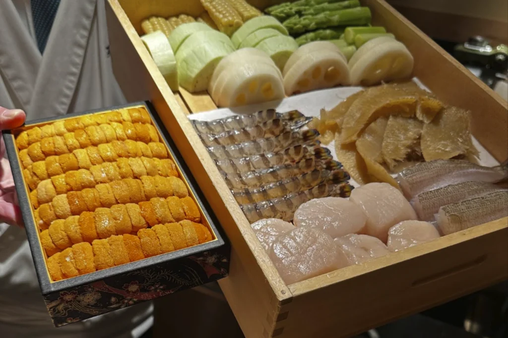A high-angle close-up of a chef presenting two wooden display boxes filled with premium raw ingredients. The smaller black box is packed with rows of bright orange Hokkaido sea urchin (uni). The larger light-wood box contains an array of fresh seasonal items: scallops, prawns, white fish fillets, lotus root, asparagus, and corn. This display showcases the "Omakase" philosophy of using the day's best market finds.