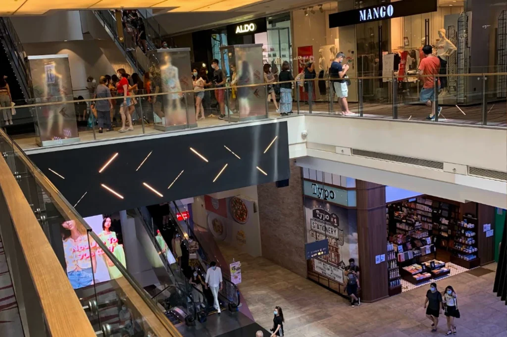This high-angle interior shot depicts a multi-level modern shopping mall bustling with people, many of whom are wearing face masks. The upper level features glass railings and storefronts for retail brands including Mango and Aldo, with shoppers walking around or standing by the edge. Escalators connect the floors, with a large, dark geometric lighting fixture visible between the levels. On the lower floor, visitors walk across the polished tile ground near a Typo stationery store, which features a rustic brick facade and well-lit shelves filled with merchandise.