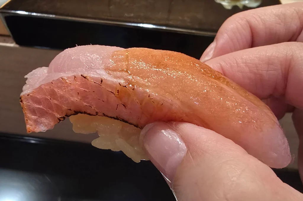 A close-up photograph captures a piece of nigiri sushi being held between a person's thumb and index finger, showcasing a thick, glistening slice of pale pink fish with a vibrant orange-tinted glaze brushed over its top surface. The fish features a distinctly seared, slightly charred bottom edge with visible grill marks, resting atop a small, compact bed of yellowish-tinted seasoned sushi rice. In the softly blurred background, a dark, glossy lacquer tray sits on a light-colored wooden counter, emphasizing the refined, intimate setting of an omakase dining experience.
