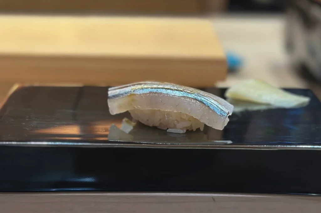 A close-up, slightly high-angle shot captures a single piece of nigiri sushi resting on a sleek, glossy black rectangular plate. The sushi features a translucent, silvery slice of fish—likely sayori (halfbeak) or a similar silver-skinned fish—with a distinct iridescent blue-green stripe running along its length, delicately draped over a hand-formed bed of seasoned sushi rice. In the soft-focused background, a piece of pickled ginger sits to the right on the plate, while a light-colored wooden sushi station board blurs into the warm, minimalist atmosphere of the restaurant.