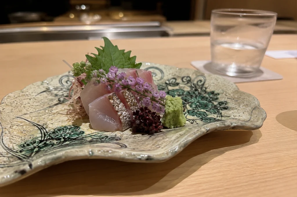 A medium shot captures an elegantly arranged plate of fresh sashimi resting on a light-colored wooden counter in a Japanese restaurant setting. The sashimi, consisting of pale pink and white fish—some slices with a textured, seared silver skin—is artfully stacked in the center of a wide, organically shaped ceramic plate. The plate features wavy, uneven edges and is decorated with intricate green and black floral patterns against a textured, sandy-beige glaze. The dish is garnished with a vibrant green shiso leaf standing upright behind the fish, a delicate stalk of tiny purple flowers draped across the front, a neat pyramid of bright green wasabi, and a small cluster of dark red seaweed. In the softly blurred background, a clear glass of water sits on a white coaster to the right, while the warm, minimalist wooden interior of the sushi bar stretches out behind the meal.