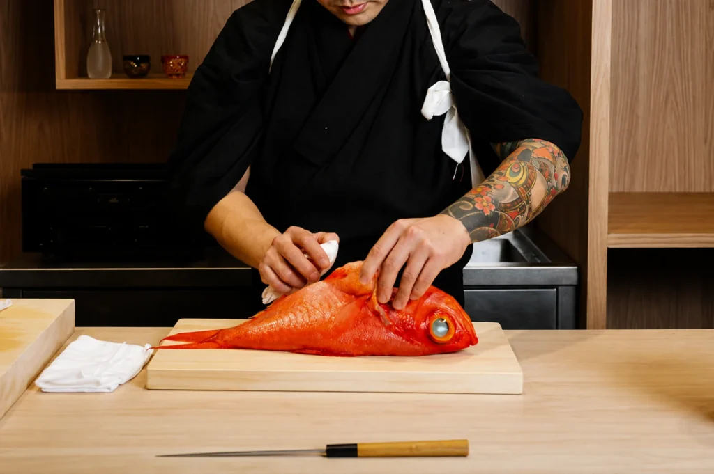 A sushi chef with visible tattoos on his arm is focused on preparing a whole, bright red fish on a wooden cutting board, using a white cloth to carefully handle the fish in a minimalist kitchen setting.