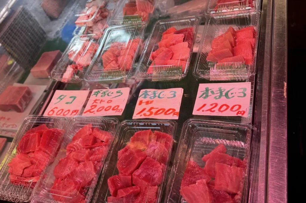 A close-up, high-angle shot of a refrigerated seafood display case containing several clear plastic trays of fresh, sliced raw tuna (maguro). The trays are arranged in two rows. The tuna varies in color from deep red Akami to pale, marbled pink Otoro (fatty tuna). Bright pink hand-written price tags in Japanese characters are placed in front of each section, showing prices ranging from 600 to 2,000 yen. The lighting is bright and cool, reflecting off the plastic packaging.