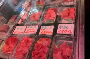 A close-up, high-angle shot of a refrigerated seafood display case containing several clear plastic trays of fresh, sliced raw tuna (maguro). The trays are arranged in two rows. The tuna varies in color from deep red Akami to pale, marbled pink Otoro (fatty tuna). Bright pink hand-written price tags in Japanese characters are placed in front of each section, showing prices ranging from 600 to 2,000 yen. The lighting is bright and cool, reflecting off the plastic packaging.