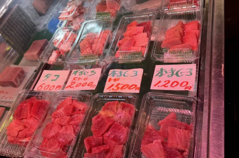A close-up, high-angle shot of a refrigerated seafood display case containing several clear plastic trays of fresh, sliced raw tuna (maguro). The trays are arranged in two rows. The tuna varies in color from deep red Akami to pale, marbled pink Otoro (fatty tuna). Bright pink hand-written price tags in Japanese characters are placed in front of each section, showing prices ranging from 600 to 2,000 yen. The lighting is bright and cool, reflecting off the plastic packaging.