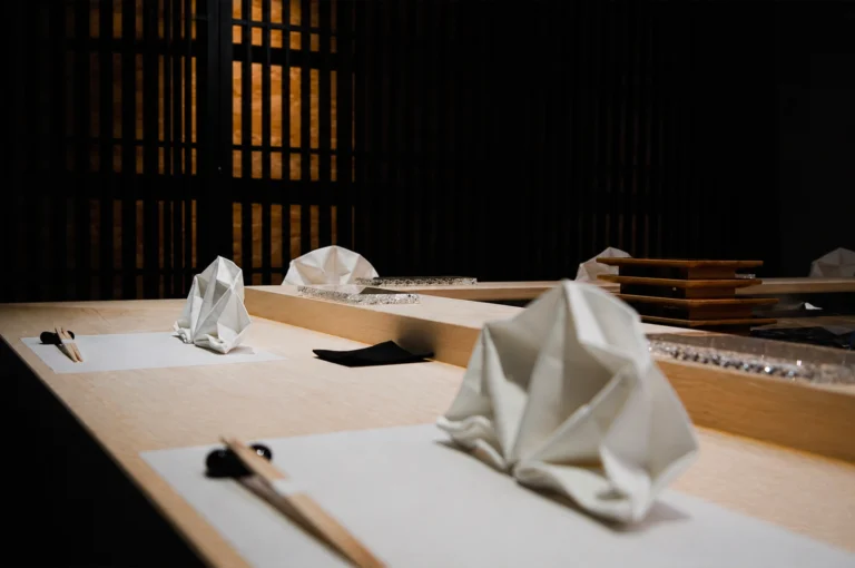 A serene, high-end sushi counter setting featuring light-colored wooden surfaces and a minimalist aesthetic. In the foreground, white cloth napkins are intricately folded into architectural, mountain-like peaks and placed on white paper placemats. Beside each placemat rests a pair of wooden chopsticks on a dark, pebble-shaped rest. The background is dominated by a dark, vertical wooden lattice screen (shoji-style), which creates a moody and intimate atmosphere through the play of light and shadow.