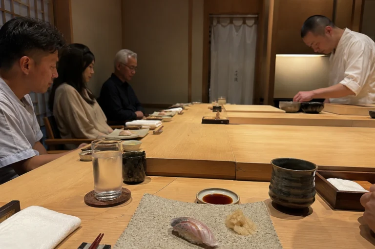 Depicts a serene and disciplined scene inside a high-end Japanese sushi restaurant, focusing on the respectful atmosphere between the diners and the chef. In the foreground, a single piece of nigiri and a mound of ginger sit on a textured ceramic plate atop a light-colored wooden counter, accompanied by a glass of water, soy sauce, and a dark tea cup. Along the left side of the long wooden bar, three diners—two men and a woman—sit in quiet anticipation, their gazes directed downward or toward the workspace. At the far end of the counter, a sushi chef with a shaved head and a white uniform is meticulously preparing food, his concentration mirroring the patient stillness of the patrons. The warm, minimalist interior, featuring traditional wooden paneling and a soft white curtain in the background, reinforces the sense of ritual and focus captured in the moment.