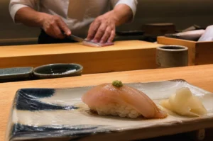 A close-up photograph features a piece of Kinmedai sushi resting on a rustic, rectangular ceramic plate with blue and white glazes. The sushi consists of a translucent slice of fish topped with a small dab of green wasabi, sitting atop a hand-pressed mound of white rice, accompanied by a pile of pickled ginger to the right. In the shallow depth-of-field background, the blurred hands of a chef in a white uniform are visible as they carefully slice a piece of fish on a light wood preparation board. The warm, professional lighting of the omakase setting highlights the glossy texture of the fish and the clean lines of the wooden counter.