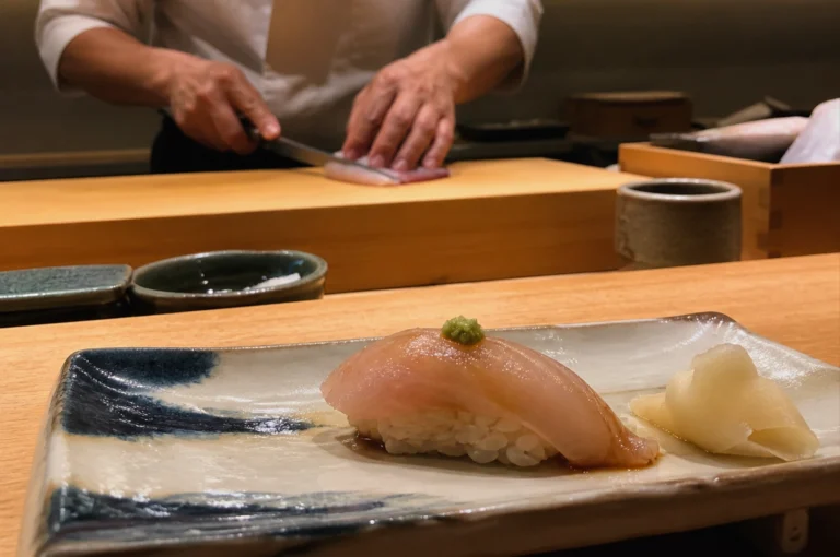 A close-up photograph features a piece of Kinmedai sushi resting on a rustic, rectangular ceramic plate with blue and white glazes. The sushi consists of a translucent slice of fish topped with a small dab of green wasabi, sitting atop a hand-pressed mound of white rice, accompanied by a pile of pickled ginger to the right. In the shallow depth-of-field background, the blurred hands of a chef in a white uniform are visible as they carefully slice a piece of fish on a light wood preparation board. The warm, professional lighting of the omakase setting highlights the glossy texture of the fish and the clean lines of the wooden counter.