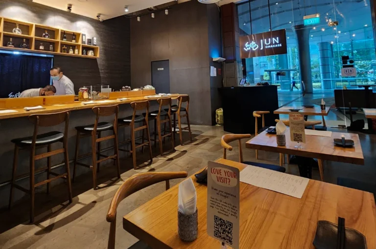 The interior of Jun Omakase restaurant in Singapore, featuring a long wooden sushi counter with high-backed stools and a separate dining area with wooden tables. A chef in a white uniform and mask is working behind the counter. Large glass windows show a modern building exterior.