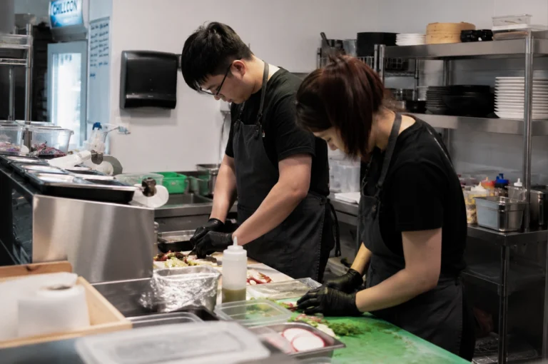 Two food service workers in a commercial kitchen are meticulously prepping ingredients at a stainless steel countertop. In the foreground, a woman with auburn hair tied back in a high ponytail leans forward over a bright green cutting board, using her gloved hands to arrange thinly sliced vegetables next to containers of sliced radishes. Beside her, a man with short black hair and glasses wears a black t-shirt and a dark grey apron over black gloves, focusing on plating or prepping ingredients on a white surface. The kitchen is brightly lit, showcasing organized stainless steel shelving units stocked with plates, bowls, and containers, while a metal food prep station with various inserts and squeeze bottles sits directly in front of them.