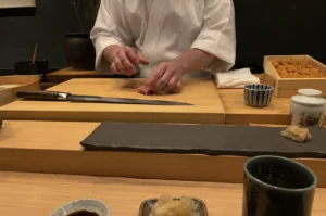 A sushi chef wearing a clean white uniform stands behind a light-colored wooden counter, meticulously preparing a piece of raw fish on a large, smooth cutting board. To the side, a long, sharp professional sushi knife rests on the board, and a wooden box filled with fresh uni is visible in the background. The minimalist setup also includes a dark, rectangular stone serving plate, small ceramic dishes containing soy sauce and pickled ginger, and a textured tea cup, all arranged in a clean, organized workspace that emphasizes high-end culinary precision.