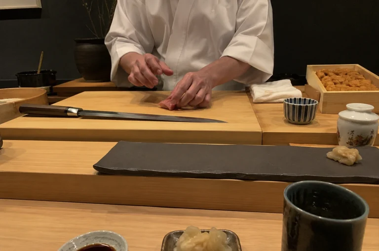 A sushi chef wearing a clean white uniform stands behind a light-colored wooden counter, meticulously preparing a piece of raw fish on a large, smooth cutting board. To the side, a long, sharp professional sushi knife rests on the board, and a wooden box filled with fresh uni is visible in the background. The minimalist setup also includes a dark, rectangular stone serving plate, small ceramic dishes containing soy sauce and pickled ginger, and a textured tea cup, all arranged in a clean, organized workspace that emphasizes high-end culinary precision.