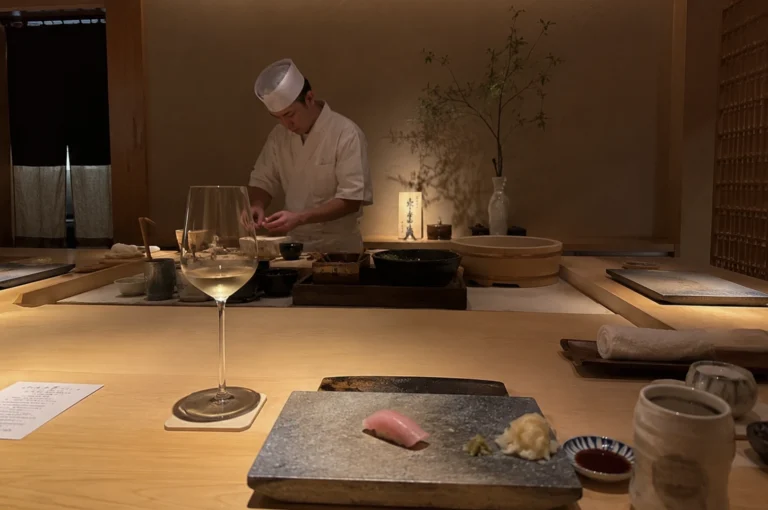 A first-person perspective from a light-colored wooden sushi counter shows a single piece of pink tuna nigiri served on a rectangular, textured gray stone plate, accompanied by a small dab of wasabi and pickled ginger. To the left sits a tall, elegant wine glass filled with white wine on a square white coaster, and to the right is a small dish of soy sauce next to a speckled ceramic tea cup and a rolled-up white hand towel. Across the immaculate counter, a sushi chef wearing a white uniform and traditional white cap is focused on preparing food, surrounded by various ceramic bowls, a wooden rice tub, and a small flower arrangement in a softly lit, minimalist restaurant setting.
