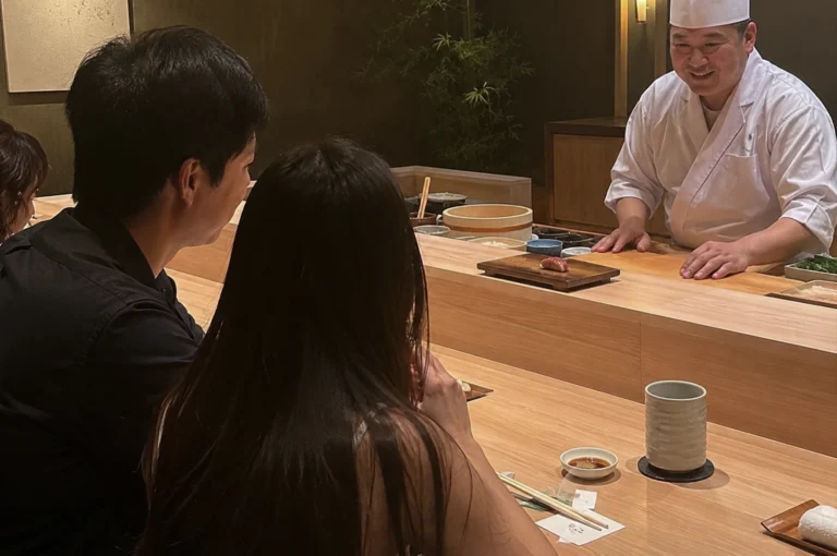 A high-angle, medium shot captures a warm interaction at a sushi bar between a male sushi chef and a dining couple. On the right, the middle-aged chef, wearing a traditional white chef’s uniform and hat, leans slightly forward with both hands on the light-colored wooden counter, smiling warmly as he prepares to speak. In front of him, a single piece of nigiri sushi sits on a small wooden serving block. Across the multi-tiered wooden counter on the left, a man in a black button-down shirt and a woman with long, straight dark brown hair sit side-by-side with their backs mostly to the camera, looking toward the chef. The woman’s hands are lightly clasped near her chin, and the profile of another diner is partially visible on the far left. The minimalist, upscale restaurant features soft, warm lighting, an earthen-toned back wall, a small bamboo plant in the background, and neatly arranged dining elements on the counter, including a ceramic teacup, chopsticks, and a small dish of soy sauce.