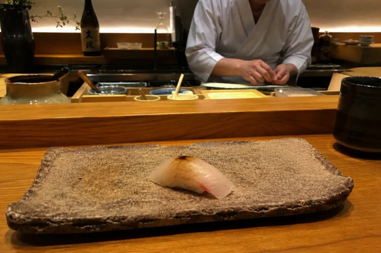 Focuses on a single piece of white fish nigiri, lightly brushed with soy sauce, centered on a textured, rectangular stone plate. The sushi sits on a polished wooden counter in a dimly lit, high-end Japanese restaurant. In the soft-focus background, a sushi chef wearing a crisp white uniform is visible from the chest down, his hands expertly shaping the next course. The scene is framed by minimalist decor, including a dark ceramic tea cup to the right and various traditional wooden prep containers and a sake bottle behind the counter, all illuminated by warm, ambient under-shelf lighting that emphasizes a serene and intimate dining atmosphere.