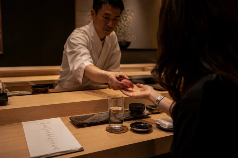 A side-angle medium shot captures a Japanese sushi chef leaning forward over a light-colored wooden counter to hand a piece of tuna nigiri directly to a female guest. The chef, wearing a clean white chef’s coat, holds the sushi delicately in his right hand, while the woman, seen from the back right foreground, extends her open palm to receive it. On the pristine wooden bar, a glass of water sits on a wooden coaster next to a ceramic soy sauce dish, a black teacup, a wet towel, and an open menu sheet lying flat.