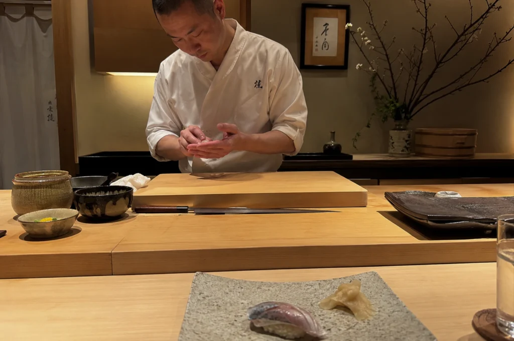Japanese sushi chef stands behind a light-colored wooden counter, meticulously shaping a piece of nigiri with his hands. He wears a traditional white chef's coat (uwagi) with a small dark insignia on the left chest. His focus is downward, directed toward his hands, where he gently presses a slice of fish onto a small bed of seasoned rice. In the foreground, a piece of finished nigiri and a small pile of pickled ginger sit on a textured, rectangular ceramic plate. The workspace is immaculately organized, featuring a long, slender yanagiba (sashimi knife) resting horizontally on the counter, alongside several small stoneware bowls and a large wooden cutting board. The background is softly lit, showcasing a minimalist aesthetic with a framed piece of calligraphy, a delicate floral arrangement in a vase, and dark wooden cabinetry, creating a serene and professional atmosphere.