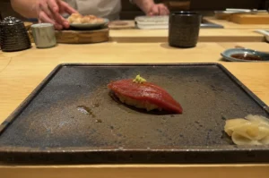 A close-up, eye-level shot captures a single piece of maguro (tuna) nigiri sushi centered on a textured, dark rectangular ceramic plate at a wooden sushi counter. The tuna has a deep red, glossy hue with delicate score marks across its surface, topped with a small dollop of bright green wasabi. In the blurred background, a sushi chef's hands are visible working with ingredients, flanked by traditional ceramic cups and small dishes on the clean wooden counter. A small cluster of pickled ginger rests on the bottom right corner of the main serving plate.