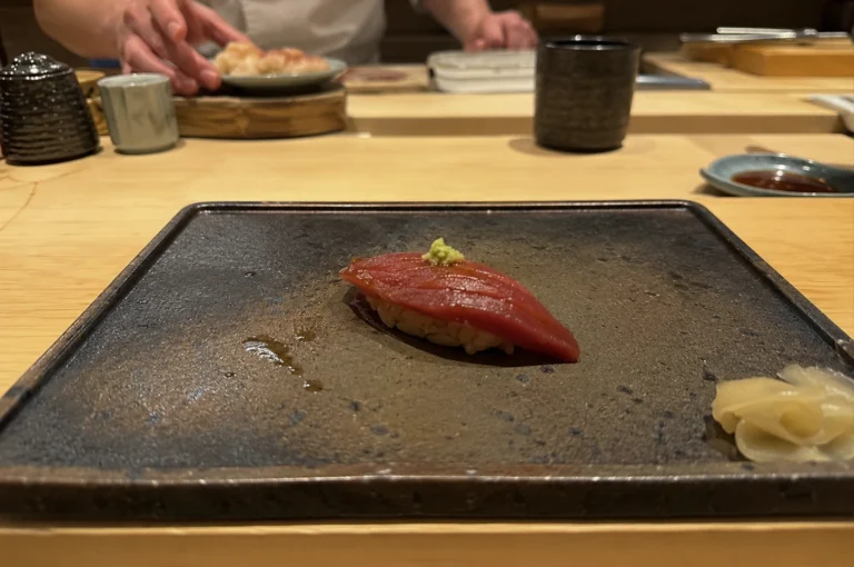 A close-up, eye-level shot captures a single piece of maguro (tuna) nigiri sushi centered on a textured, dark rectangular ceramic plate at a wooden sushi counter. The tuna has a deep red, glossy hue with delicate score marks across its surface, topped with a small dollop of bright green wasabi. In the blurred background, a sushi chef's hands are visible working with ingredients, flanked by traditional ceramic cups and small dishes on the clean wooden counter. A small cluster of pickled ginger rests on the bottom right corner of the main serving plate.