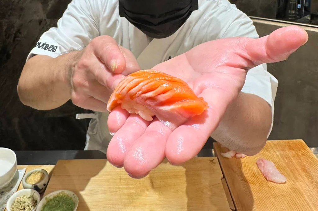 A first-person perspective shot highlights a sushi chef presenting a freshly crafted piece of salmon nigiri held directly in his open palm toward the diner. The bright orange, scored salmon fillet glistens with glaze over a small bed of rice, set above a wooden prep board lined with small bowls of condiments.
