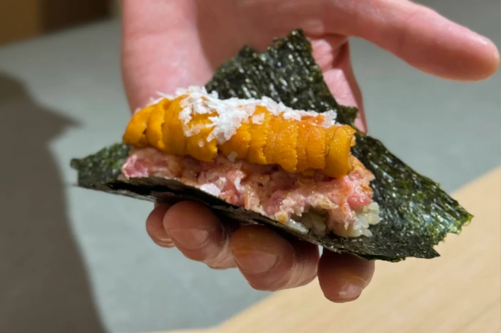 A hand-held perspective showcases a luxurious "Uni and Negitoro" handroll wrapped in a crisp, dark green sheet of nori seaweed. The roll is generously layered with a base of seasoned rice, a thick spread of chopped fatty tuna (negitoro), and topped with several vibrant orange lobes of fresh sea urchin (uni). A generous sprinkling of flaky white sea salt is visible on top of the sea urchin, emphasizing the premium quality and textural contrast of the ingredients.