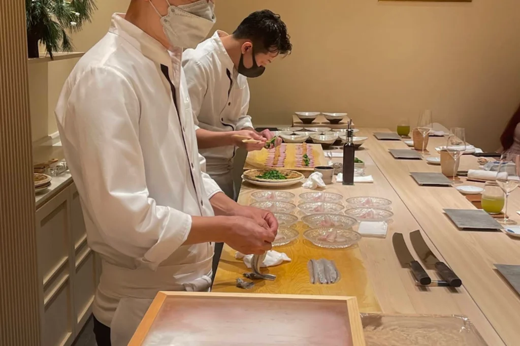Two professional sushi chefs, both wearing white uniforms and protective face masks, are meticulously preparing multiple servings at a long, light-colored wooden sushi counter. The chef in the foreground is focused on carefully plating delicate portions of silver-skinned fish onto a series of clear glass plates, while the second chef in the background is precisely arranging ingredients on a wooden board. The counter is lined with various dining essentials, including elegant glassware filled with green tea, small condiment bottles, and professional-grade sushi knives resting on the wooden surface.