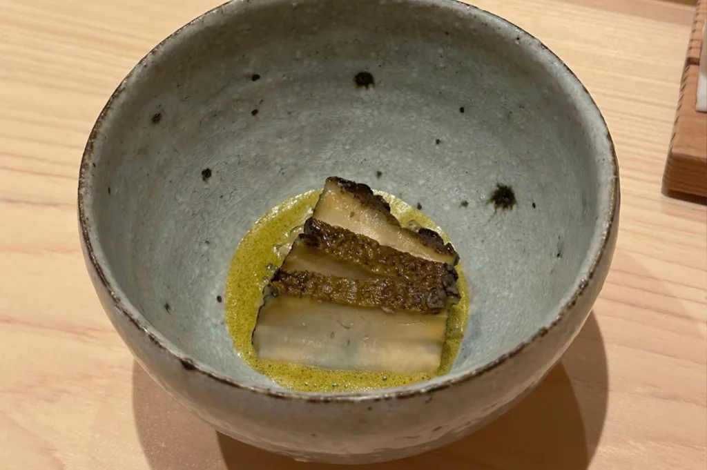 A close-up, top-down view shows three thick, tender slices of braised abalone resting in a shallow pool of vibrant green liver sauce inside a rustic, light blue-grey ceramic bowl. The abalone pieces feature a dark, textured outer edge and a smooth, cream-colored interior, perfectly glazed and glistening under the light. The bowl itself has a beautiful crackle glaze finish with subtle dark speckles, and the entire dish is presented on a clean, light-grained wooden tabletop.