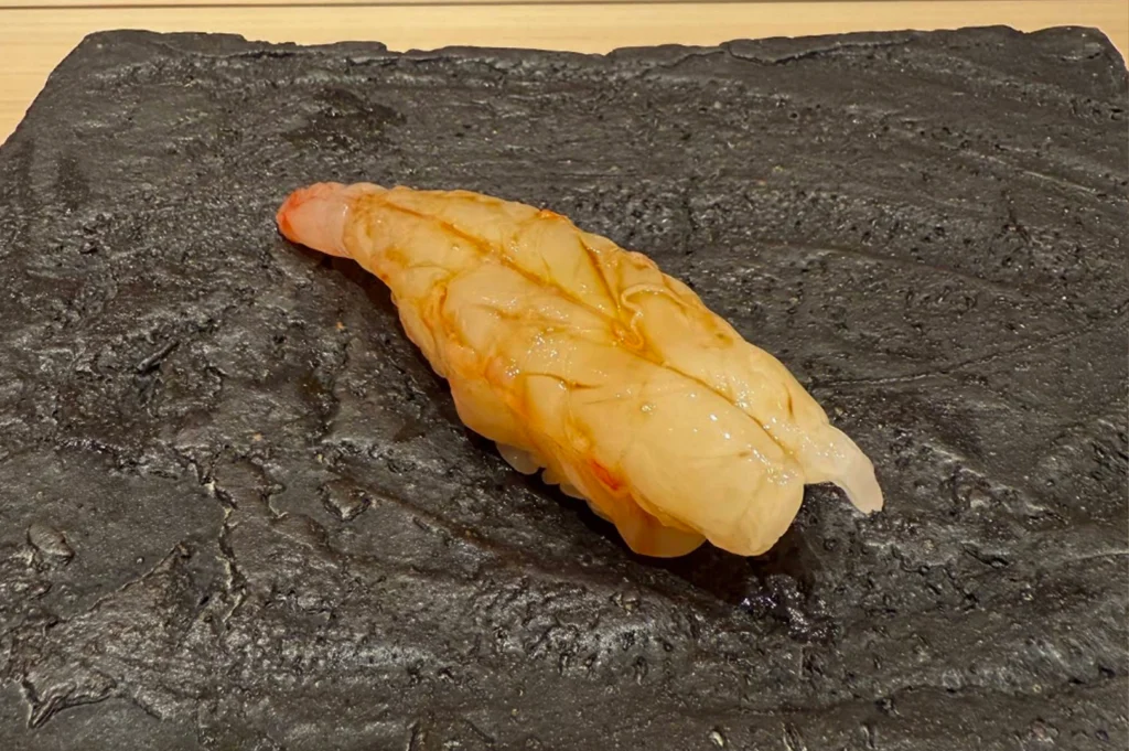 A single piece of premium Botan Ebi (spotted prawn) nigiri is presented on a large, textured black stone plate that has a rugged, matte finish. The translucent prawn is expertly butterflied and draped over a small bed of seasoned sushi rice, featuring a delicate orange hue and a glossy sheen from a light brush of soy glaze. The dark, uneven surface of the artisanal stone plate provides a dramatic and sophisticated contrast to the delicate, pale colors of the fresh seafood.