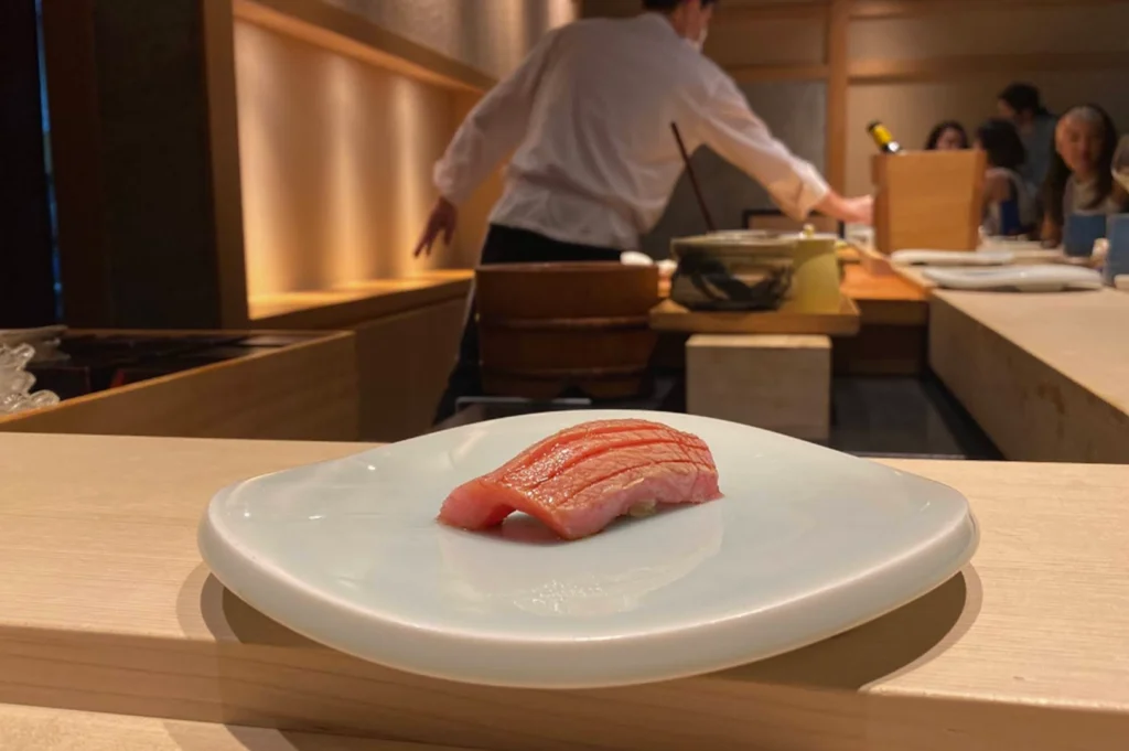 A single piece of premium otoro nigiri, featuring fatty tuna with distinct, scored lines across its pink surface, is presented on a minimalist, square white ceramic plate. In the background, the blurred setting of an upscale omakase restaurant is visible, showing a chef in a white uniform preparing dishes behind a wooden counter while diners sit in the distance. The lighting is warm and focused, highlighting the delicate texture of the fish against the clean lines of the sushi bar.