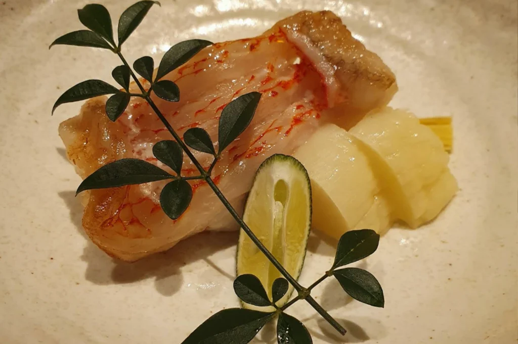 A beautifully presented cooked fish dish rests in a shallow ceramic bowl, featuring a succulent fillet with visible red skin markings. It is accompanied by several slices of pale, tender bamboo shoots and a small wedge of green citrus lime. A delicate green branch with dark leaves is laid across the fish for an artistic touch, emphasizing the fresh, seasonal ingredients used in a traditional Japanese omakase course.