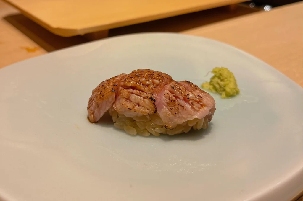 A close-up of a piece of aburi (torched) nigiri sushi sits on a light-colored, square plate. The fish topping is lightly charred on the surface to bring out its oils and smoky flavor, resting on a small bed of seasoned sushi rice. A tiny mound of fresh green wasabi is placed neatly to the side on the plate, all set against the soft, warm tones of a wooden sushi counter.