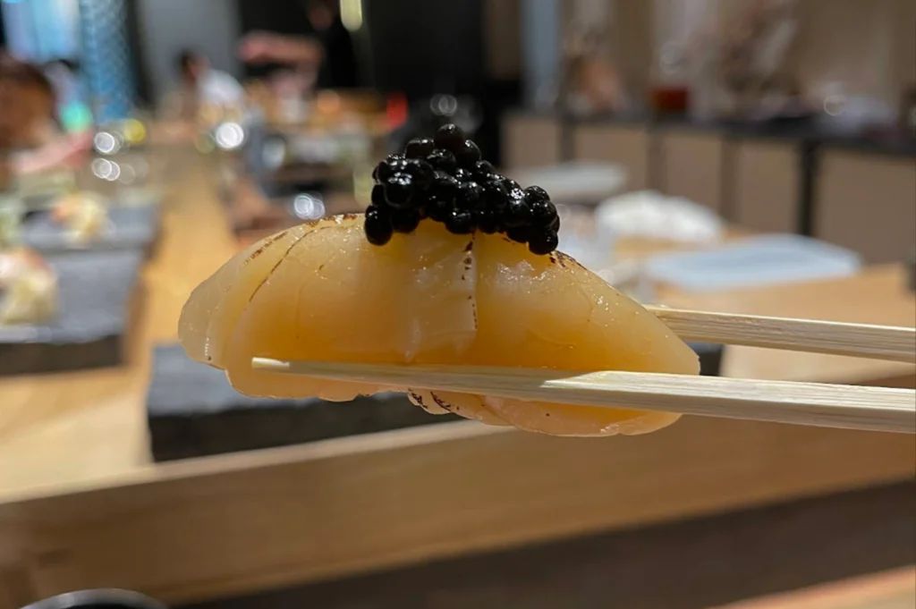 A pair of wooden chopsticks holds a piece of scallop nigiri sushi, which is topped with a generous portion of glistening black caviar. The scallop is translucent and slightly torched at the edges, resting on a compact base of rice. The shot is a close-up with a shallow depth of field, making the luxurious ingredients stand out against the blurred background of the restaurant interior.