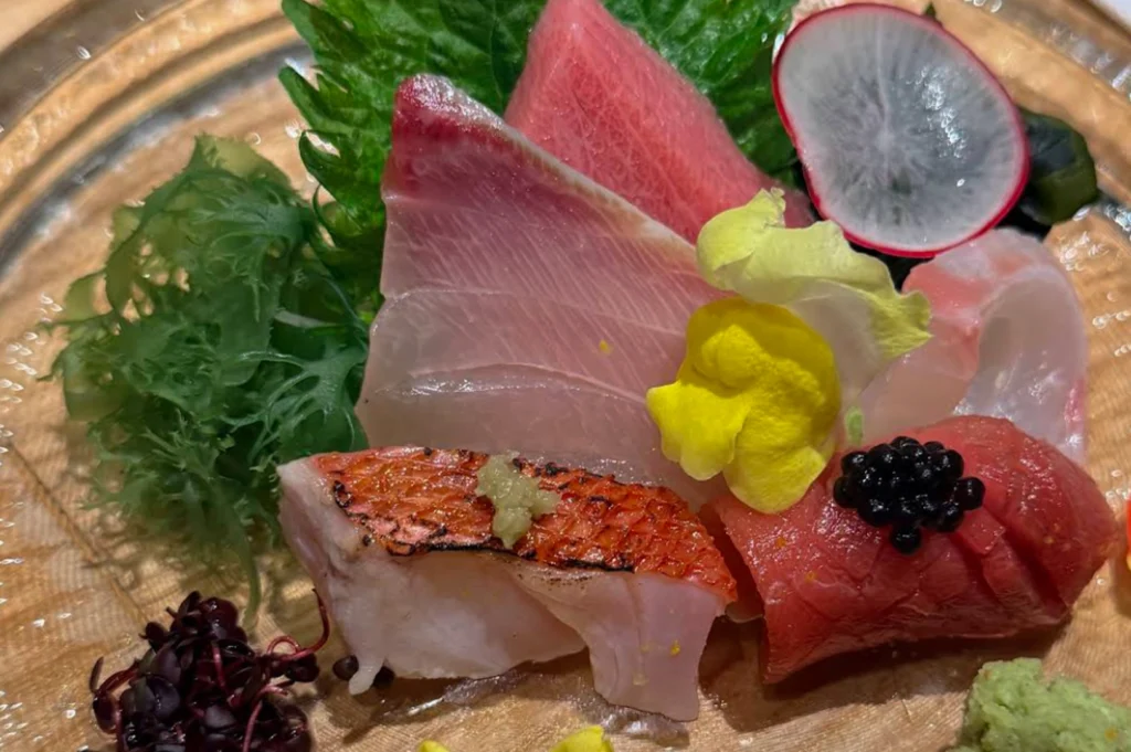 An vibrant assortment of fresh sashimi is artistically arranged on a clear glass plate. The selection includes thick slices of deep red tuna, translucent white fish, and a piece of fish with bright red skin topped with a touch of ginger. The dish is garnished with a green shiso leaf, a thin slice of radish, a bright yellow edible flower, a small cluster of black caviar, and a mound of fresh wasabi, creating a colorful and high-end presentation.