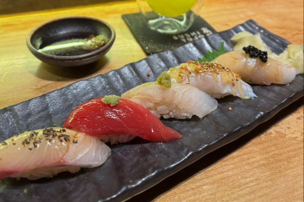 A row of five different types of nigiri sushi is neatly arranged on a long, textured black rectangular platter. The sushi varieties include white fish with different seasonings, a bright red lean tuna, and a piece topped with black caviar. In the background, a small dark bowl of soy sauce with wasabi and a glass of green tea sit on the wooden counter, ready for the diner.