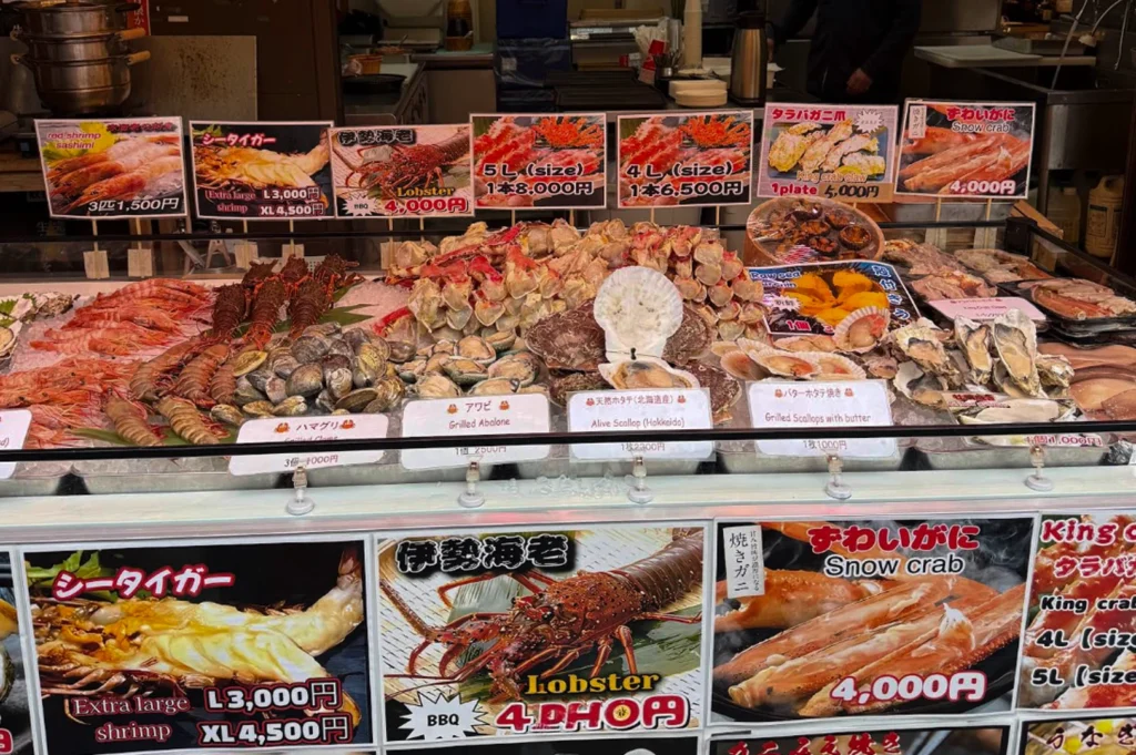 A wide shot of an outdoor seafood stall featuring a tiered display of shellfish on ice. The display includes large red shrimp, spiny lobsters, massive king crab legs, abalone, and scallops in their shells. Above the seafood, large colorful placards show pictures of the cooked versions of these items with prices: King Crab legs are listed at 6,500 to 8,000 yen, and grilled lobsters at 4,000 yen. The bottom of the display features large bilingual signs in Japanese and English for "Sea Tiger" shrimp and "Snow Crab."