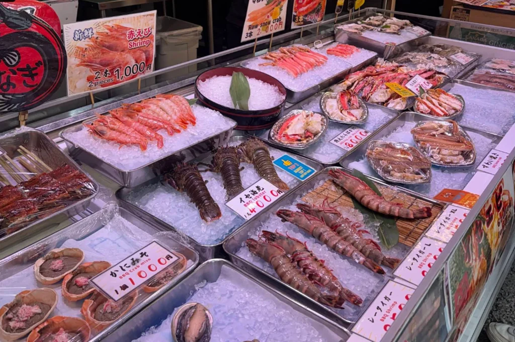 This vibrant photograph captures a densely packed refrigerated display case filled with a high-end variety of fresh seafood arranged on beds of crushed ice. Trays feature bright red shrimp, large spiny lobsters, various types of crab legs, and open crab shells filled with "kani miso" (crab brain paste). Each item is accompanied by small price tags in Japanese, such as "1,000 Yen" for shrimp or "4,000 Yen" for lobster. The lighting is bright and clean, emphasizing the glossy textures of the raw shellfish and the meticulous presentation typical of a Japanese fish market.